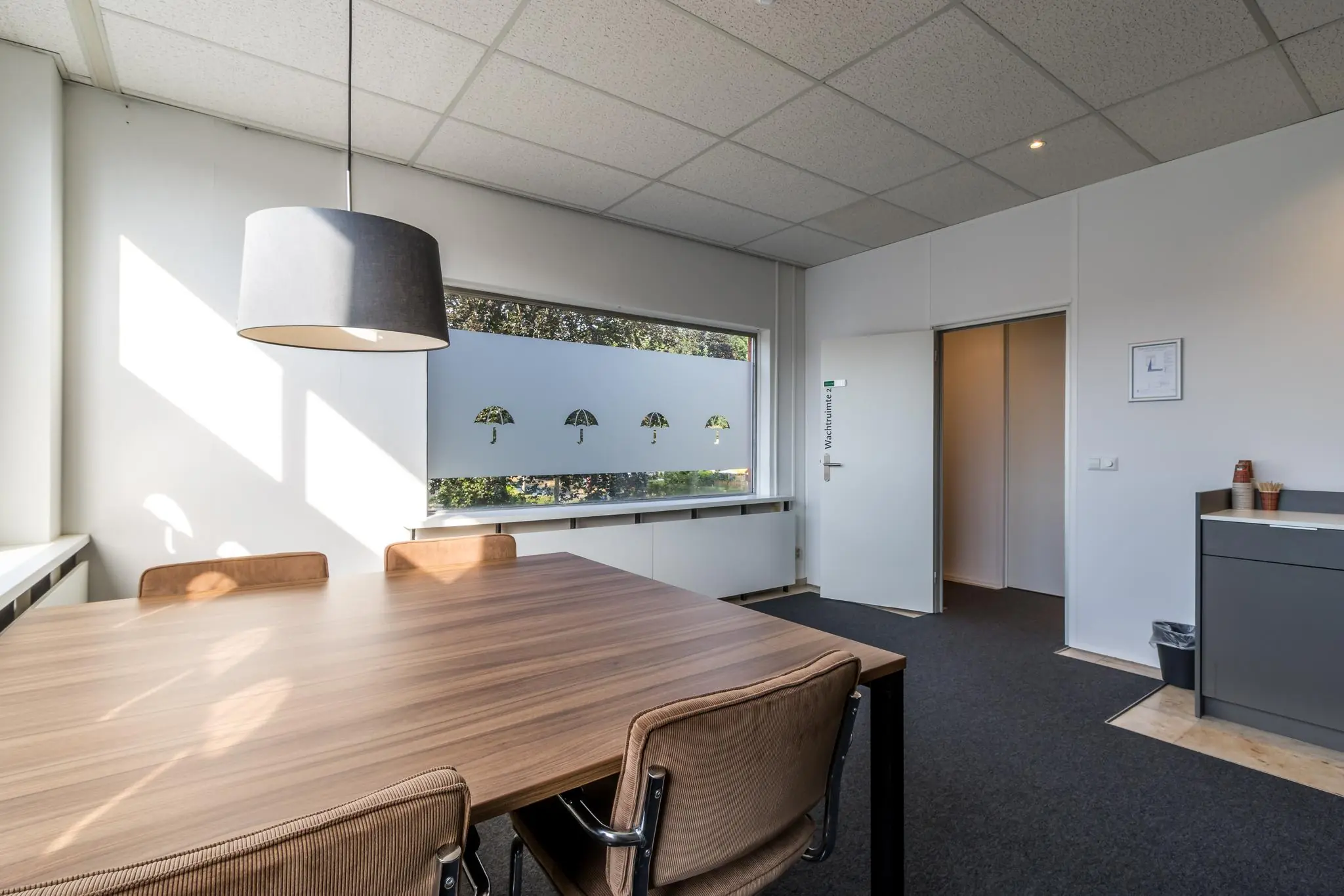 Vergaderruimte aan de Jan van Eycklaan met houten tafel, bruine stoelen, grote hanglamp en raamdecoratie met paraplu-motief.